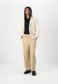 Lichtbeige blazer over een zwart shirt, gecombineerd met losse beige broek en zwarte schoenen. Stevige stof en op maat gemaakte ontwerp met zakken.