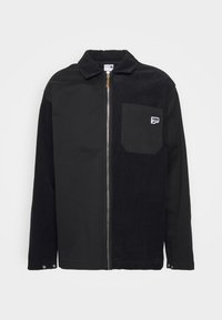 Giacca nera con una texture in cotone a coste, chiusura con zip e colletto a punta. Caratterizzata da una tasca sul petto sinistro con etichetta del marchio. Bottoni ai polsini.