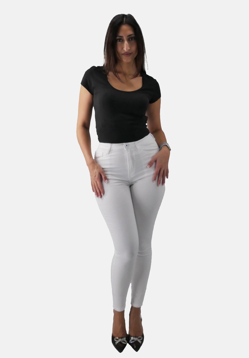 Maglietta nera aderente abbinata a jeans skinny bianchi a vita alta. La modella indossa tacchi neri con accenti argentati, in piedi con le mani sui fianchi.