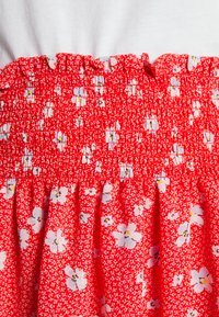 Saia vermelha com franzido, apresentando um padrão floral branco, com textura franzida e uma cintura com babados. O material de algodão proporciona um acabamento suave.