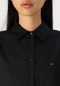 Zwart overhemd met kraag en knoopsluiting, met een klein multikleurig logo aan de linkerkant. Glad stof en slim fit.