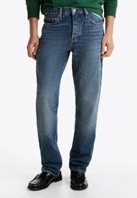 Blå denim jeans med lige ben, let falmning, fem lomme-design og standard knappelukning. Båret med sorte sko.