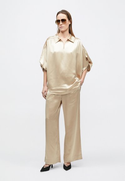 Žena nosi béžovú blúzku z voľného saténu a široké nohavice, tmavé slnečné okuliare a čierne špicaté lodičky, stojaca s jednou rukou v vrecku.