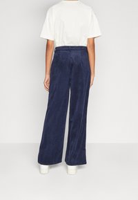 Široké kordové kalhoty v tmavě modré barvě s texturovaným povrchem, spárované s krémovým tričkem, nošeny s bílými botami.