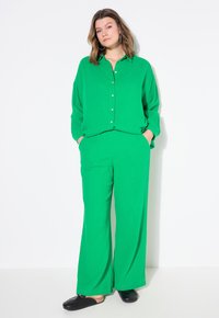 Ensemble deux pièces vert composé d'une chemise à boutons à coupe décontractée et de pantalons à jambe large. Fabriqué en tissu texturé ; modèle portant des chaussures noires.