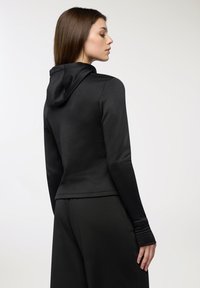 Felpa nera attillata con maniche lunghe, caratterizzata da un cappuccio a profilo basso e aperture per il pollice sui polsini, realizzata in tessuto liscio ed elastico.