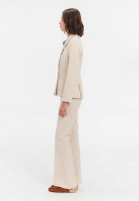 Costume beige ajusté avec une veste cintrée et un pantalon évasé, présentant un tissu texturé et un motif rayé subtil. Chaussures accent marron.