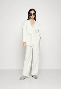 Moteris, vilkinti baltą kostiumą ir leopardų rašto nėrinius, papuošta dideliais juodais akinių rėmeliais ir ilgomis kabančiomis auskarais.