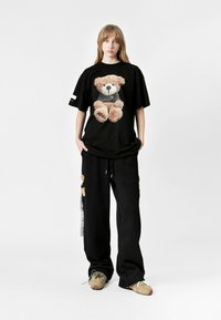 Una camiseta negra de gran tamaño presenta un gráfico texturizado de un oso; combinada con pantalones negros holgados adornados con acentos de oso; el modelo lleva zapatillas beige.