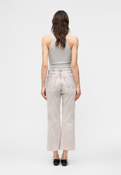 Γυναίκα με κυματιστά καστανά μαλλιά που φοράει γκρι αμάνικη μπλούζα, φωτεινά τζιν με ίσια μπατζάκια και μαύρα επίπεδα παπούτσια, στέκεται γυρισμένη με την πλάτη σε λευκό φόντο.