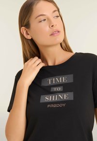 Camiseta negra con un estampado decorativo que incluye las palabras "TIME TO SHINE" y "FREDDY," acentuadas con tachuelas metálicas. Manga corta, corte relajado.