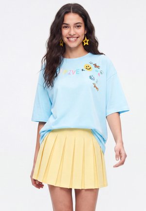 Ung kvinde smiler, iført en lyseblå oversized t-shirt med farverige prints og et gult plisseret skørt, mod en enkel hvid baggrund.