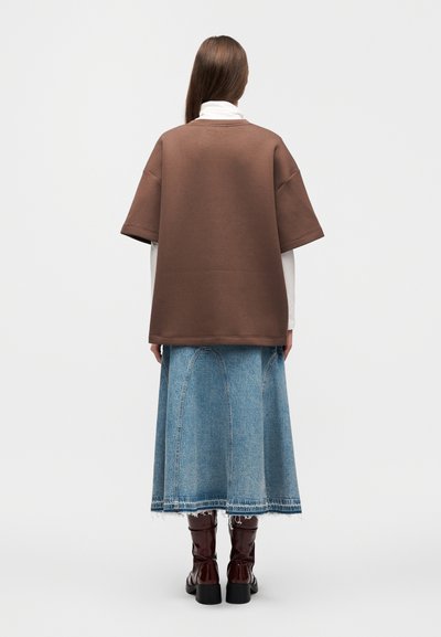 Bruine oversized sweater gecombineerd met een lange blauwe spijkerrok. Model draagt bruine kniehoge laarzen. Eenvoudige achtergrond.