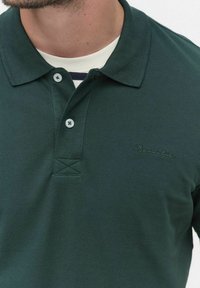 Mörkgrön polo skjorta i texturerad tyg, har en krage, tre-knapp placket och broderad logotyp i en ljusare nyans.