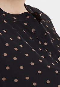 Černá blůza s kulatými béžovými puntíky. Obsahuje kulatý výstřih a légu na knoflíky se zlatými knoflíky. Měkká textilie.