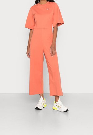 Combinaison corail à manches courtes et jambes larges, en coton. Associée à des baskets Nike blanches et vert néon, ornées d'un logo swoosh.