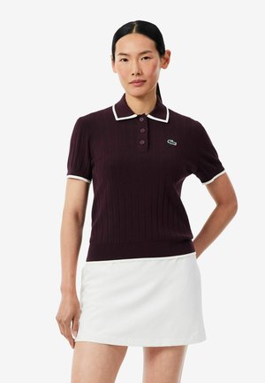 Femme portant un polo manches courtes violet foncé avec un liseré blanc et une jupe blanche, debout avec une main derrière le dos.