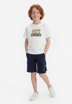 Παιδί με κόκκινα μαλλιά που φοράει λευκό μπλουζάκι με γραφικό σχέδιο αρκούδας, ναυτικό cargo σορτς με τσεπάκι λογότυπο και λευκά αθλητικά παπούτσια, στέκεται με τα χέρια στις τσέπες.