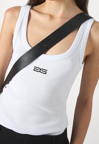 Kvinna iförd en vit ribbad linne med en svart logotyp och ett brett svart läderband som korsar hennes bröst diagonalt.