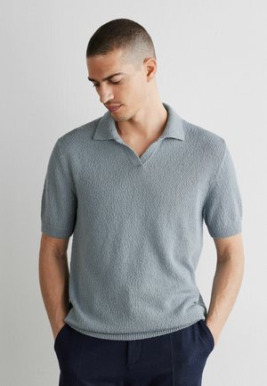 Man met kort haar, gekleed in een lichtblauw polohemd met korte mouwen en structuur, en een donkere broek, kijkt naar beneden met handen in de zakken.