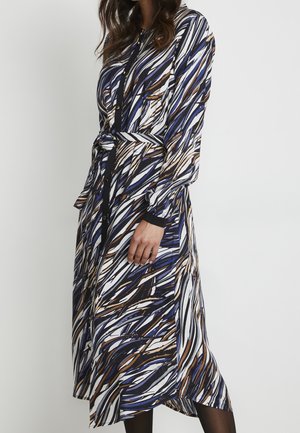 Kvinde iført en langærmet, knappet kjole med et abstrakt penselstrøgsmønster i blå, brun, sort og hvid samt et matchende bælte i taljen.