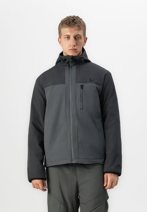 Young man wearing a two-tone dark gray zip-up jacket with a hood and matching cargo pants, standing against a plain white background.