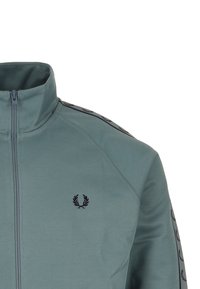 Chaqueta de chándal azul-verde con cremallera y logo de laurel negro en el pecho, además de rayas negras con patrón a lo largo de las mangas.