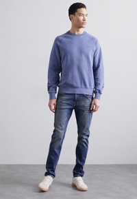 Blå hettegenser med ribbestrikkede mansjetter og nederkant, i kombinasjon med mørke denimjeans og hvite joggesko, stående på en grå gulv mot en lys bakgrunn.