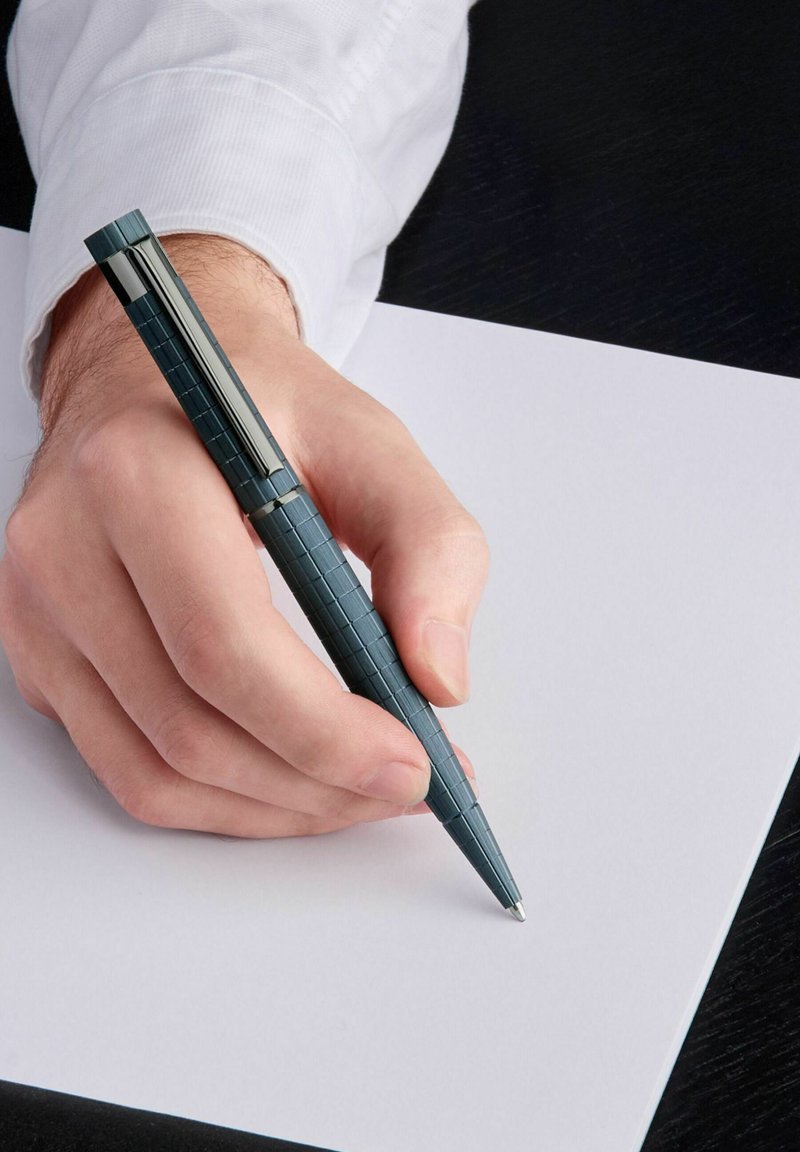 Stylo bleu foncé avec une prise texturée et nervurée, tenu au-dessus d'une feuille blanche vide. Accent de clip argenté près du haut.