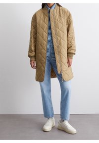 Osoba ubrana w beżową pikowaną długą kurtkę na niebieską kurtkę dżinsową i jasnoniebieskie dżinsy, w białych tenisówkach na szarym podłożu.