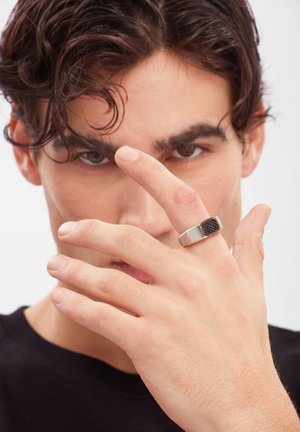 Junger Mann mit dunklem Haar trägt einen silbernen Ring mit einem schwarzen strukturierten Quadrat am Mittelfinger, der teilweise sein konzentriertes Gesicht bedeckt.