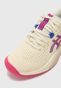 Chaussure de sport beige clair avec tige perforée, semelle en caoutchouc rose, accents bleus et roses sur les lacets et le logo. Matériau texturé et détails de design.
