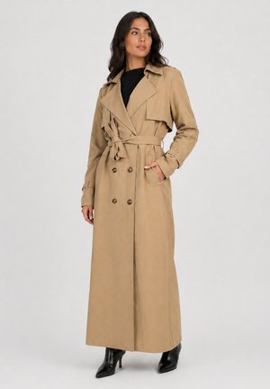 Kvinde stående iført en lang beige dobbeltradet trenchcoat med bælte, sort top og sorte støvler med hæl på en ensfarvet baggrund.
