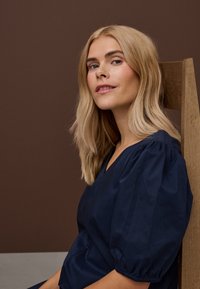 Plavolasa ženska s ramensko dolgimi lasmi, oblečena v temno modro bluzo z nabranimi rokavi, sedi bočno na lesenem stolu pred rjavim ozadjem.