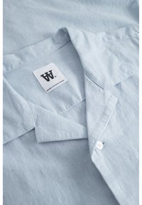 Chemise en lin bleu clair avec un col, dotée d'un bouton blanc et d'une étiquette avec du texte noir. Le tissu a une surface douce et texturée.