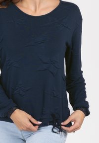 Blusa de manga larga azul marino con acentos texturizados en capas y un cordón en el dobladillo. Tiene un escote redondeado y puños con un sutil brillo.