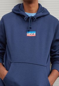 Felpa con cappuccio blu navy con coulisse, con logo Levi's rosso e blu sulla tasca frontale. Materiale in cotone, texture morbida.
