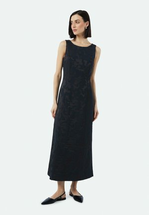 Ärmelloses, bodenlanges schwarzes Kleid mit einem dezenten floralen Muster, gefertigt aus strukturiertem Stoff. Kombiniert mit schwarzen Slingback-Ballerinas.
