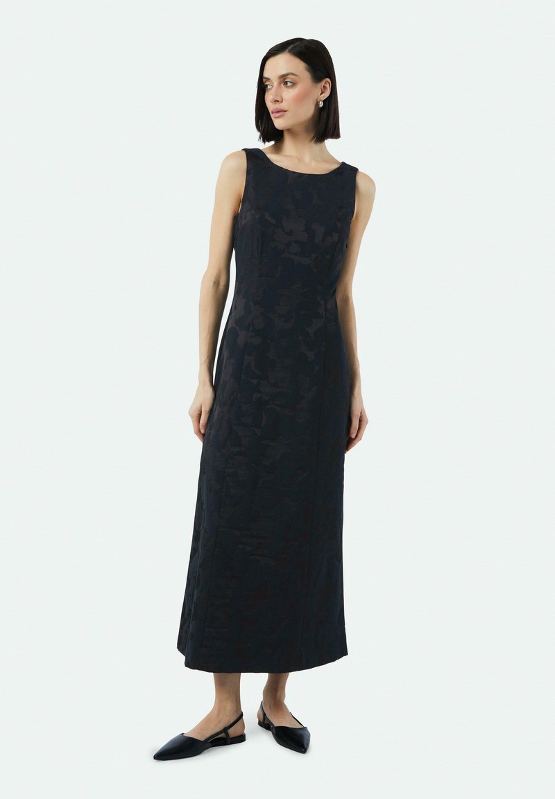 Mouwloze, vloerlange zwarte jurk met een subtiel bloemenpatroon, gemaakt van textuurstof. Gepaard met zwarte slingback ballerina's.