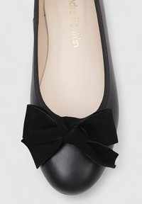 Ballerine plate en cuir noir avec un bout rond, ornée d'un nœud en velours noir sur le devant. Texture lisse et design minimaliste. Marque en or à l'intérieur.