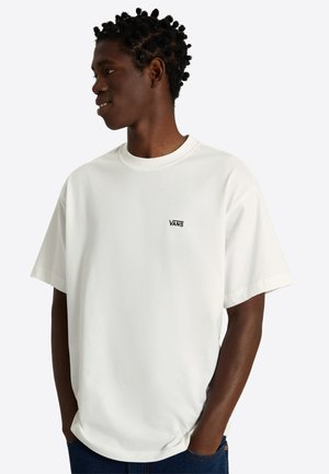 Jonge man met kort gedraaid haar, draagt een wit Vans T-shirt en blauwe spijkerbroek, kijkt opzij met handen in de zakken tegen een effen achtergrond.