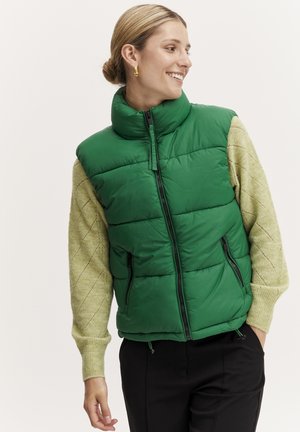 Groene gewatteerde vest met hoge kraag, ritssluiting aan de voorkant en zijkanten, gedragen over een lichtgroene gebreide trui met een ruitpatroon.