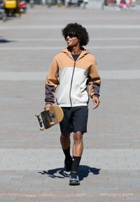 Chaqueta con capucha en beige, crema y gris; pantalones cortos negros; patineta en mano; lleva calcetas negras y zapatillas deportivas; caminando por un camino pavimentado.