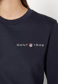 Personne portant une chemise bleu marine avec le texte "GANT", un logo bouclier avec des étoiles et des rayures, et "1949" imprimé sur la poitrine près de l'encolure.