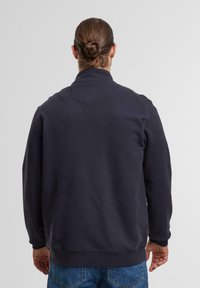 Dunkelblaues Sweatshirt mit hohem Kragen, strukturiertem Stoff und gerippten Bündchen. Sichtbare Nähte und eine lässige Passform. Getragen mit hellblauen Jeans.
