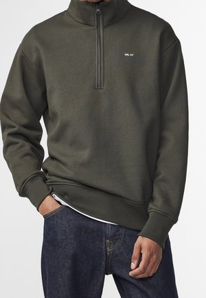 Mann trägt dunkelolivfarbenes Quarter-Zip Sweatshirt mit „NN.07“-Logo auf der Brust und blaue Jeans, Hand in der Tasche vor einfarbigem Hintergrund.