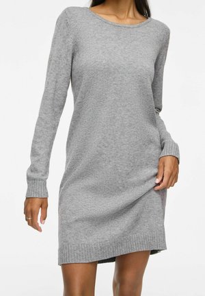Donna che indossa un abito in maglia grigio chiaro a maniche lunghe con polsini e orlo a coste, tenendo leggermente l'abito con una mano.