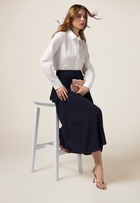 Chemise blanche à manches longues avec des broderies, associée à une jupe bleu marine foncé. Les accessoires comprennent une pochette rose gold et des sandales à talons.