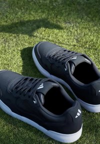 Črne športne čevlje z gladko teksturo, ki imajo perforirane detajle, bele podplate in tri bele črte na straneh.