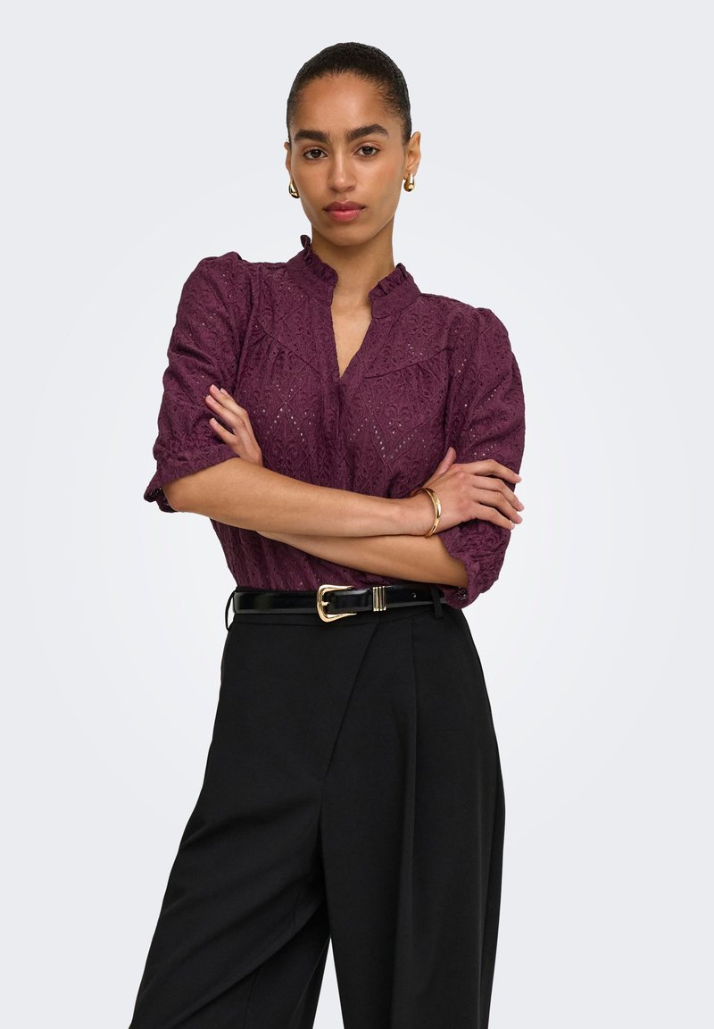 Femme aux cheveux plaqués en arrière porte un chemisier violet texturé et un pantalon taille haute noir avec une ceinture noire, debout les bras croisés.